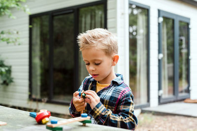 Little boy in a checkered shirt plays with a wooden constructor outdoors.