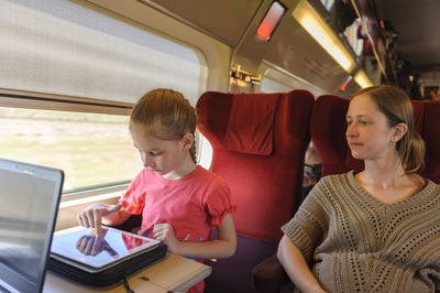 Woman looking at girl using digital tablet in train