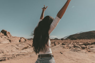 Rear view of young woman with arms raised standing at desert against sky during sunny day