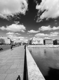 People walking on footpath by river against cloudy sky