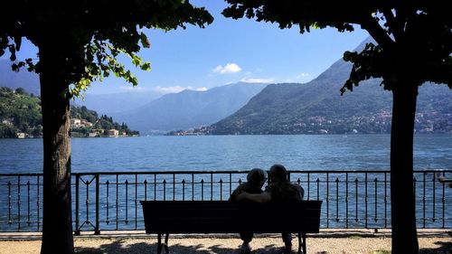 Rear view of couple sitting on bench by lake against sky