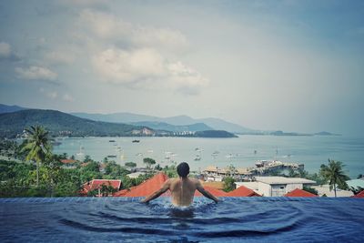 Rear view of shirtless man in infinity pool looking at sea against sky