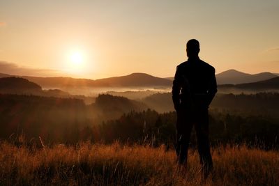 Tall hiker stand on meadow with golden stalks of grass and watch over misty and foggy valley 