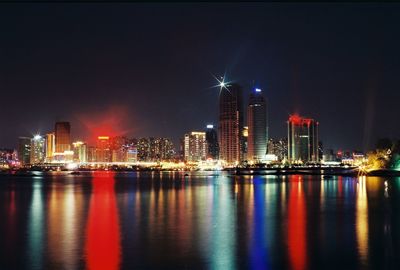 Illuminated modern buildings reflecting on river against sky at night