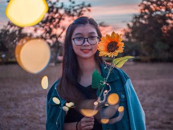 Portrait of smiling young woman holding sunflower with illuminated string lights during sunset