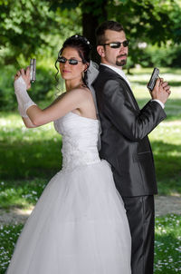 Portrait of couple in wedding dress holding guns while standing back to back at park