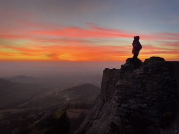 Silhouette of a woman standing on top of a tall cliff and watching the sunset