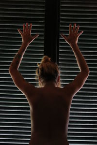 Rear view of naked woman with arms raised standing by window