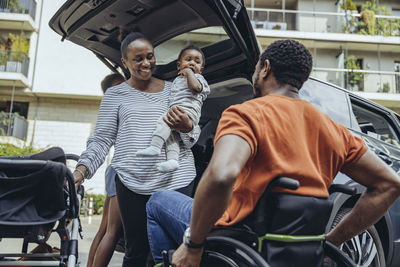 Mother carrying son looking at father in wheelchair by car