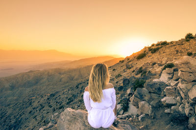 Rear view of woman sitting on rock against sky during sunset