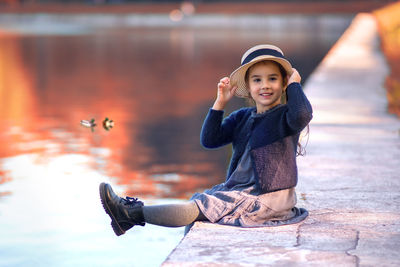 A cheerful little girl sits on the shore of a pond,
