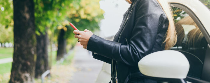 Trendy blonde girl using smartphone leaning against a car. teen using mobile phone chat with friends