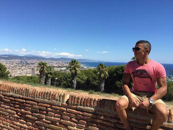 Man sitting on retaining wall against sky