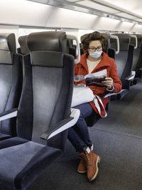 Woman in red duffle coat and protective mask reads magazine in suburban train. new normal.
