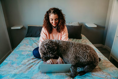 Smiling woman using laptop by dog on bed at home