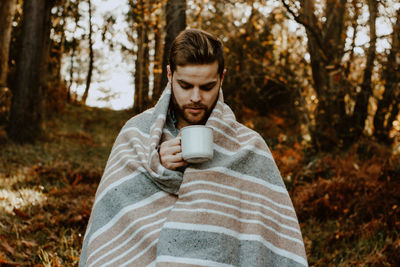 Young man wrapped in blanket having coffee in forest