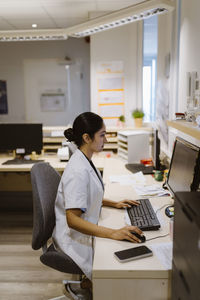 Side view of female healthcare worker working on computer sitting in clinic