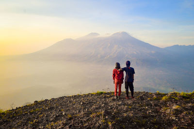 Rear view of couple standing on mountain against sky during sunset