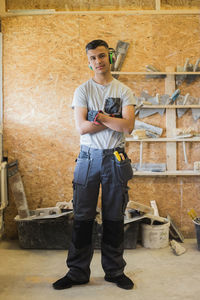 Full length portrait of confident teenage trainee standing with arms crossed against wall at workshop