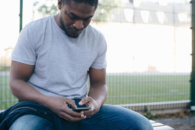 Young man using smart phone while sitting on bench against fence