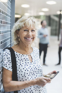 Portrait of happy businesswoman holding digital tablet while leaning on wall in office