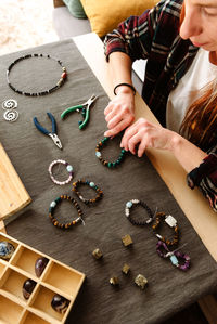 From above cropped unrecognizable female traveler sitting at table in truck and creating handmade bracelets during trip