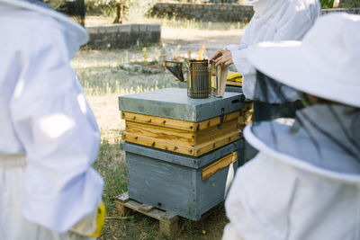 Unrecognizable crop beekeepers on protective costumes using bee smoker in apiary in summer