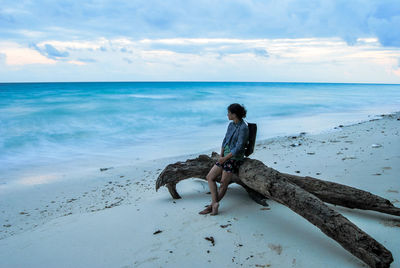 Young woman sitting on log at beach against cloudy sky during sunset