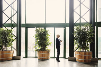 Businesswoman using smart phone while standing by potted plants in office