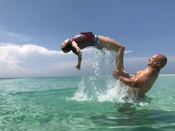 Side view of girl jumping from shirtless father in sea against sky