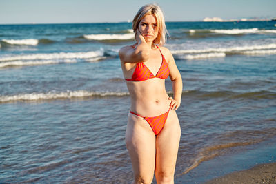 Blond woman in red bikini holding hand on waist and touching neck while standing on beach near waving sea on summer weekend day