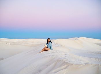 Full length of woman sitting on sand dune against sky during sunset