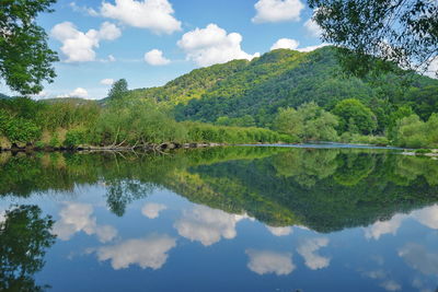 A calm lake in wich the green hills and trees, the blue sky and the white clouds reflect.