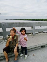 Grandmother and granddaughter eating ice cream on bridge against sky during sunset
