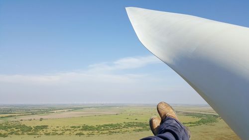 Low section of person beside part of wind turbine against cloudy sky