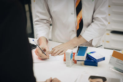 Midsection of female pharmacist writing on prescription paper with senior customer standing at checkout in medical store