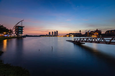 Illuminated bridge over river by buildings against sky during sunset