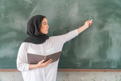 Teacher wearing traditional clothing writing on blackboard while standing in classroom