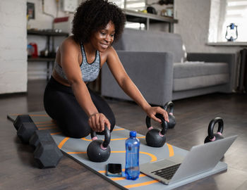 Happy ethnic sportswoman with kettlebells and laptop