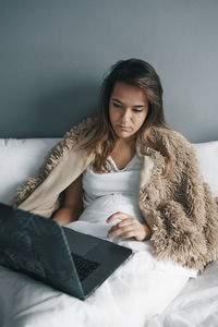 Sick young woman using laptop on bed against wall at home