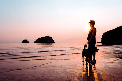 Silhouette senior woman with dog standing at beach against sky during sunset