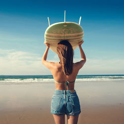 Rear view of young woman holding surfboard while standing at beach against sky