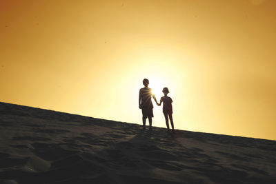 Silhouette siblings standing on sand against sky during sunset