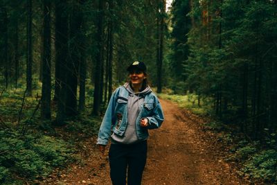 Portrait of smiling woman wearing cap walking on dirt road amidst forest