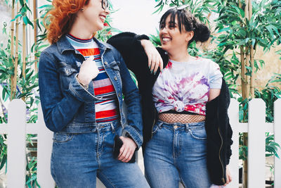 Smiling hipster female friends standing against fence