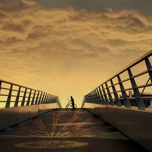 Mid distance view of man with bicycle on footbridge against sky during sunset