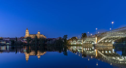 Illuminated buildings by river against clear blue sky at night