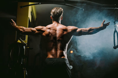 Rear view of shirtless man with arms outstretched amidst smoke in gym