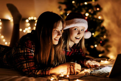 Two charming sisters lie on the bed with a laptop on christmas or new year's eve 