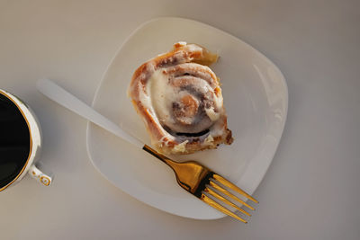 Cinnamon bun on a white plate and elegant fork on the white table, top view. 